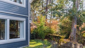 A house with a dark siding exterior, a visible window, and a garden with trees and shrubs. - Creekside Hideout for Quiet Souls Condo A (Nanoose Bay)