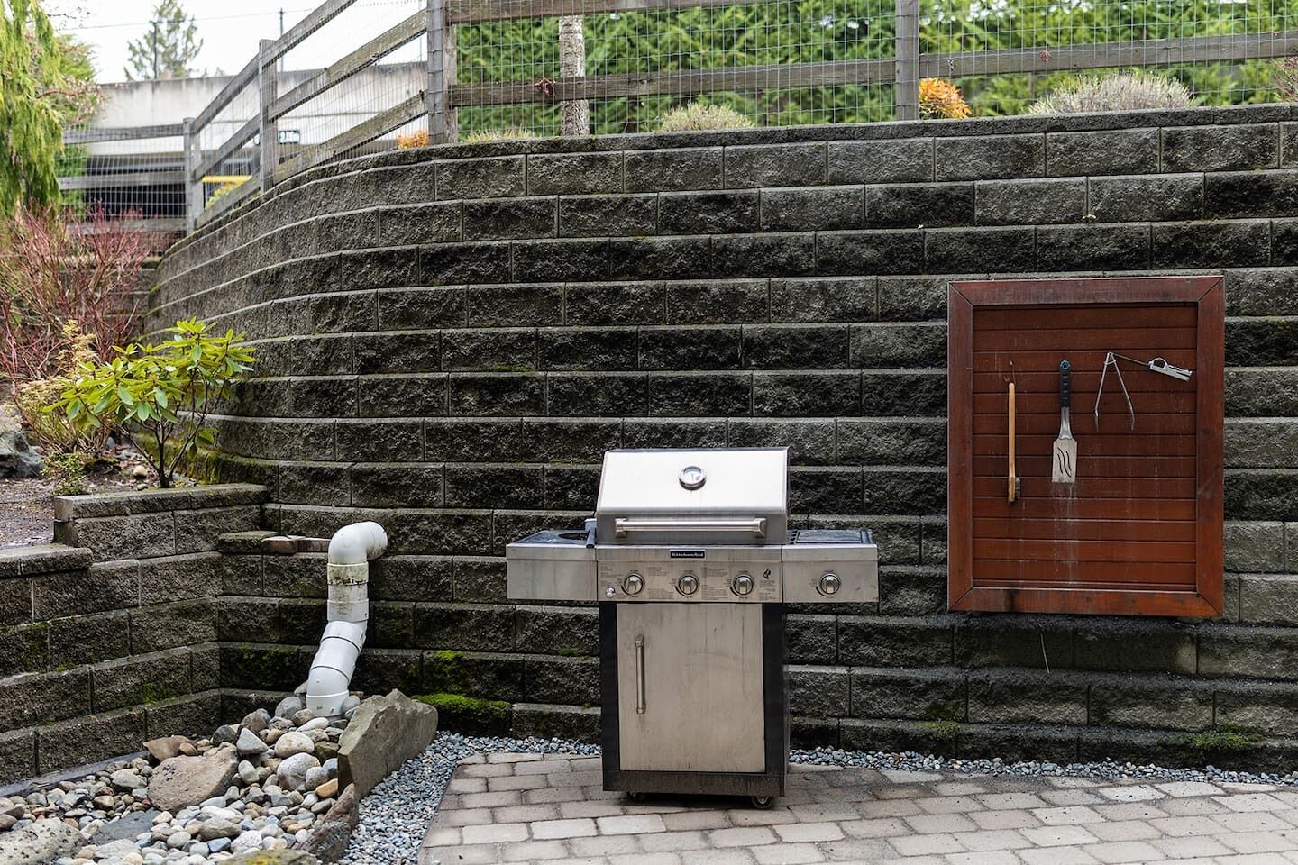 A barbecue grill with a wooden door and a metal handle, set against a stone wall with a metal fence in the background.