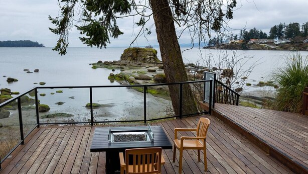 A wooden deck with a glass-top table and two chairs, overlooking a rocky shoreline and calm water.