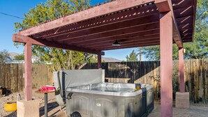 Een hottub onder een houten pergola met een stenen terras, omringd door een houten omheining en bomen.