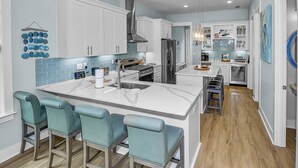 A modern kitchen with white cabinets, a stainless steel refrigerator, a built-in oven, and a large island with bar stools. - Community Pool/3min to Beach/Screened Patio/2 Lofts/Fire Pit/Foosball&Pingpong/*Golf Cart/Sleeps 16 (Santa Rosa Beach)