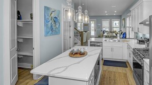 A modern kitchen with white cabinetry, a central island, pendant lighting, and a dining area with a sofa and table. - Community Pool/3min to Beach/Screened Patio/2 Lofts/Fire Pit/Foosball&Pingpong/*Golf Cart/Sleeps 16 (Santa Rosa Beach)