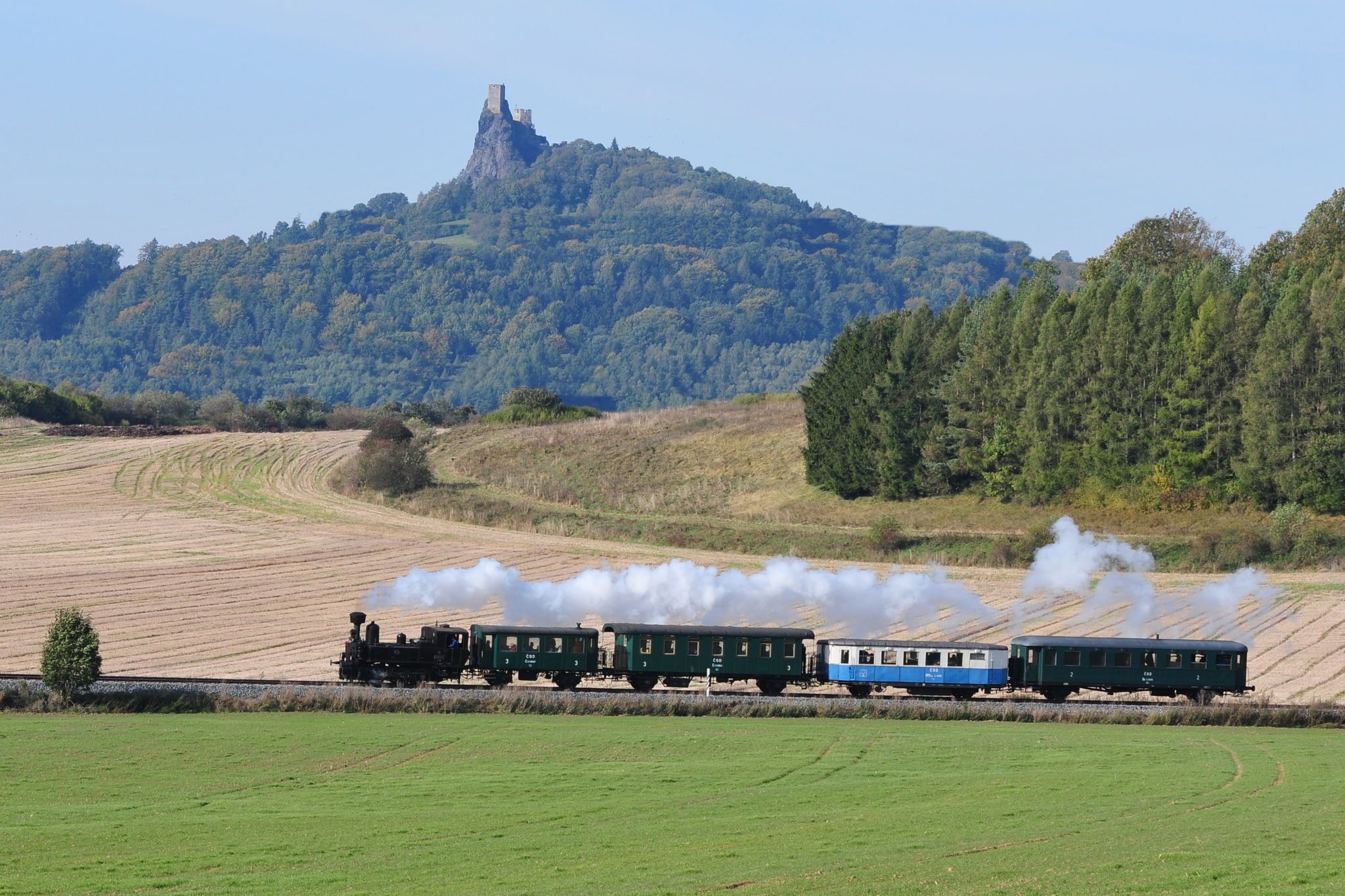 A steam train with multiple carriages moving through a rural landscape with fields and a hill with a castle in the background.