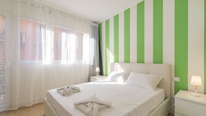 A hotel room with a bed, white curtains, a green and white striped wall, a bedside lamp, and a nightstand.