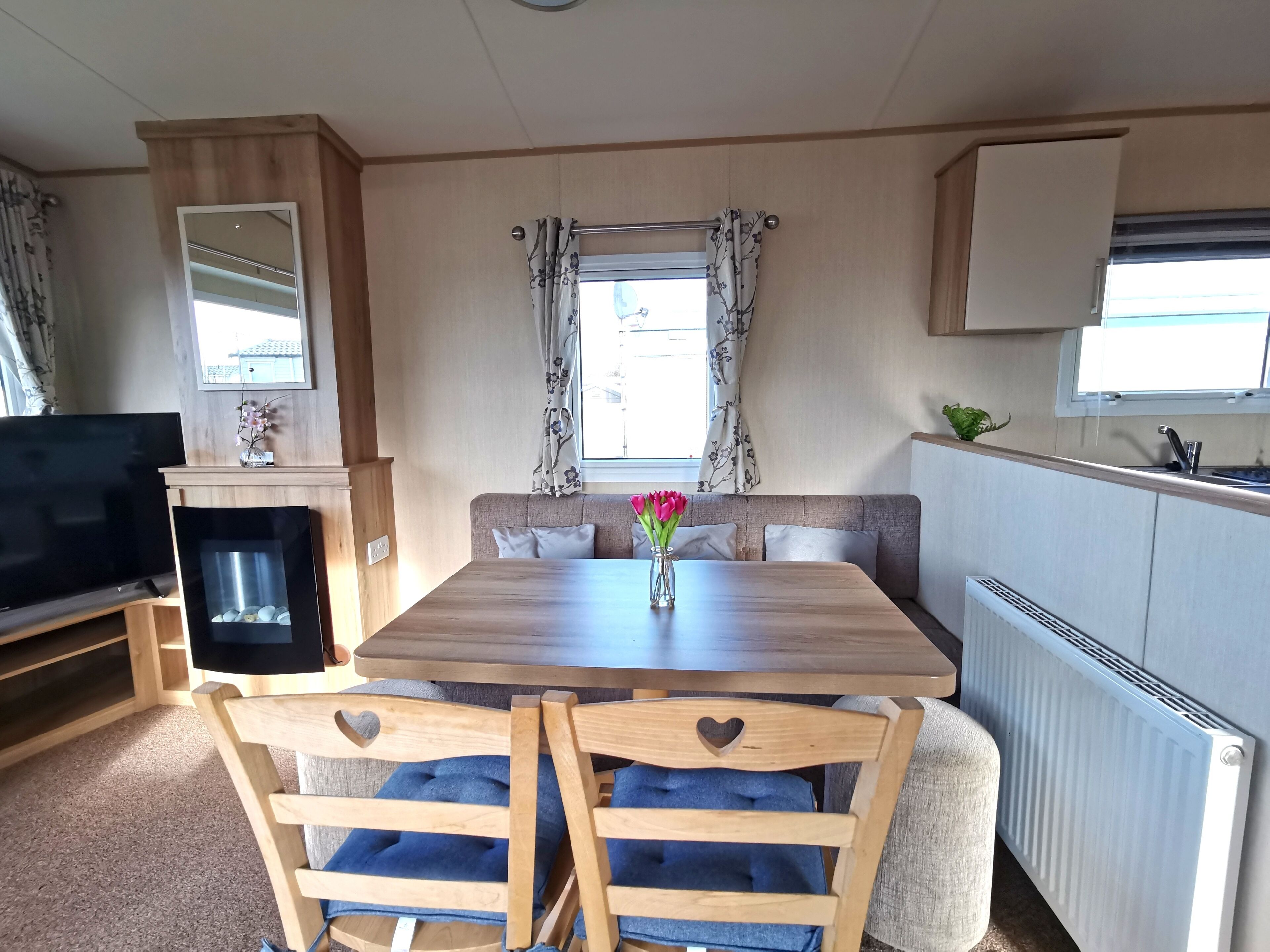 Interior acolhedor de uma RV com uma mesa de jantar de madeira, cadeiras com almofadas azuis, uma televisão e uma pequena cozinha.
