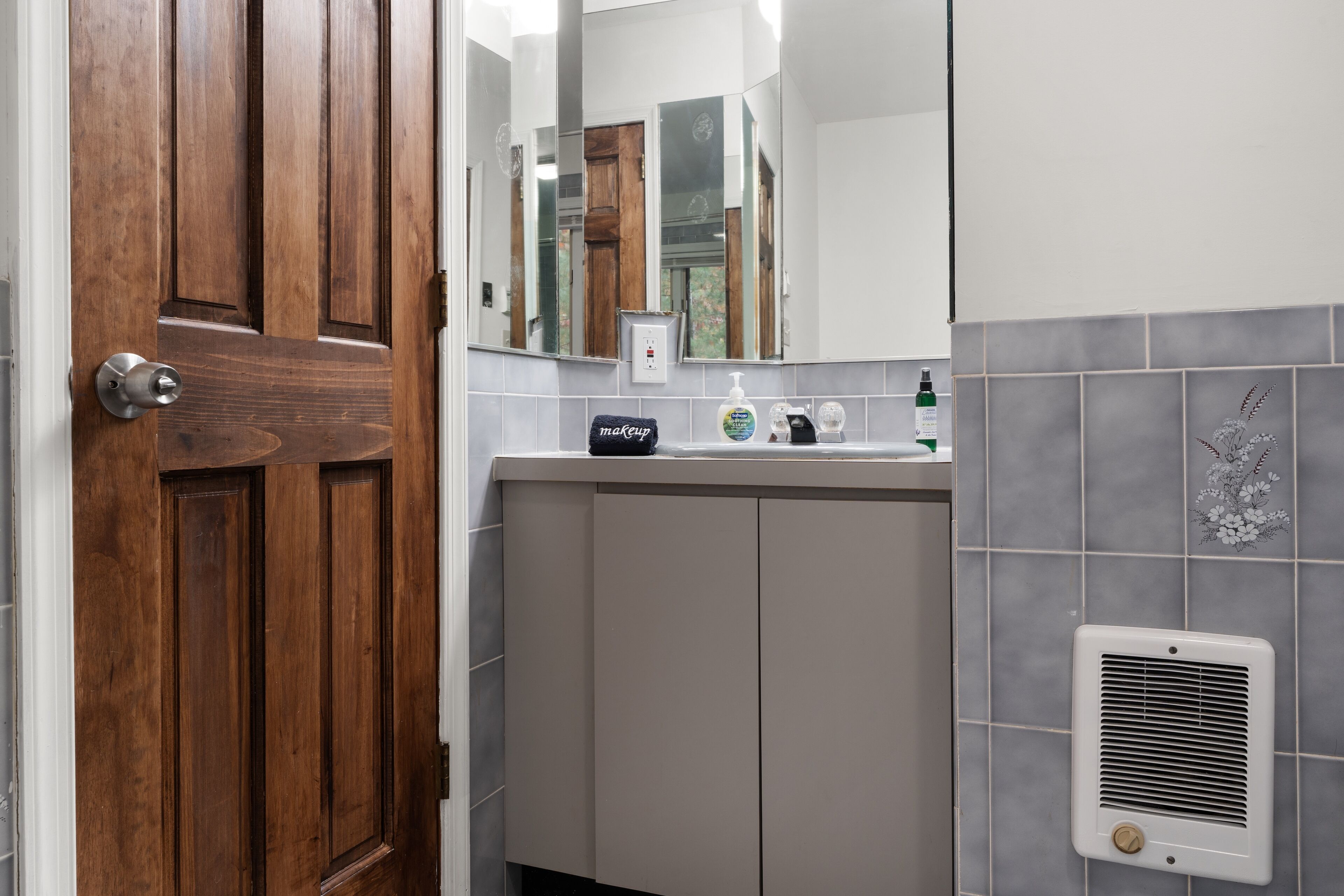 A bathroom with a wooden door, a mirror, a sink with a makeup dispenser, and a vent on the wall.