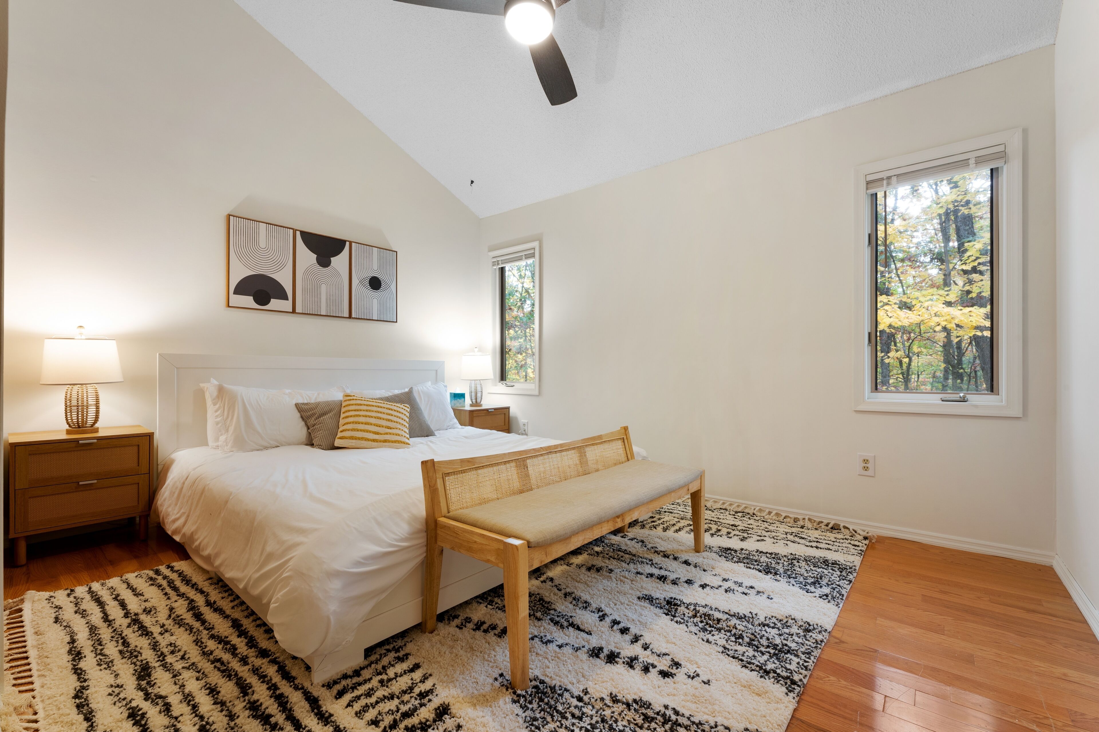 Un dormitorio con cama, mesitas de noche, un banco y un amplio ventanal con vistas a árboles.
