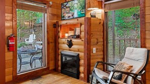 Ein gemütliches Blockhaus-Interieur mit Kamin, einem Wand-Fernseher und einem Sessel mit Kissen.