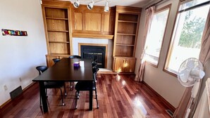 Une pièce spacieuse dotée d’un plancher en bois, d’une table à manger avec des chaises, d’étagères intégrées en bois, d’une cheminée et d’une fenêtre donnant sur les arbres.