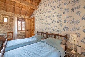 A room with a stone wall, wooden ceiling beams, a bed with blue bedding, a wooden nightstand with a lamp, and a floral wallpaper pattern.