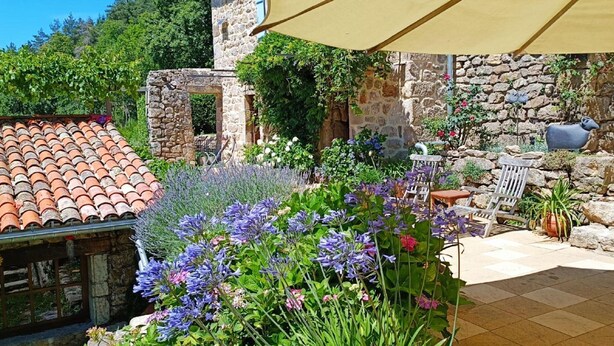 Un bâtiment en pierre, surmonté d’un toit en tuiles, entouré d’une végétation luxuriante et de fleurs.