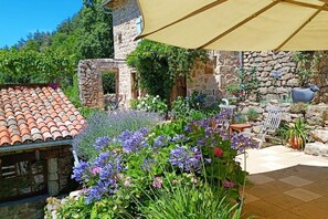 Un bâtiment en pierre, surmonté d’un toit en tuiles, entouré d’une végétation luxuriante et de fleurs.