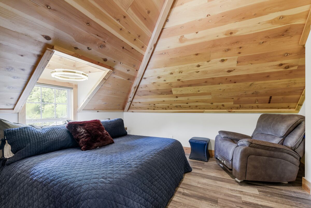 Een slaapkamer met een schuine kap, een bed met een gewatteerde spreidek, een lederen fauteuil en een klein krukje.