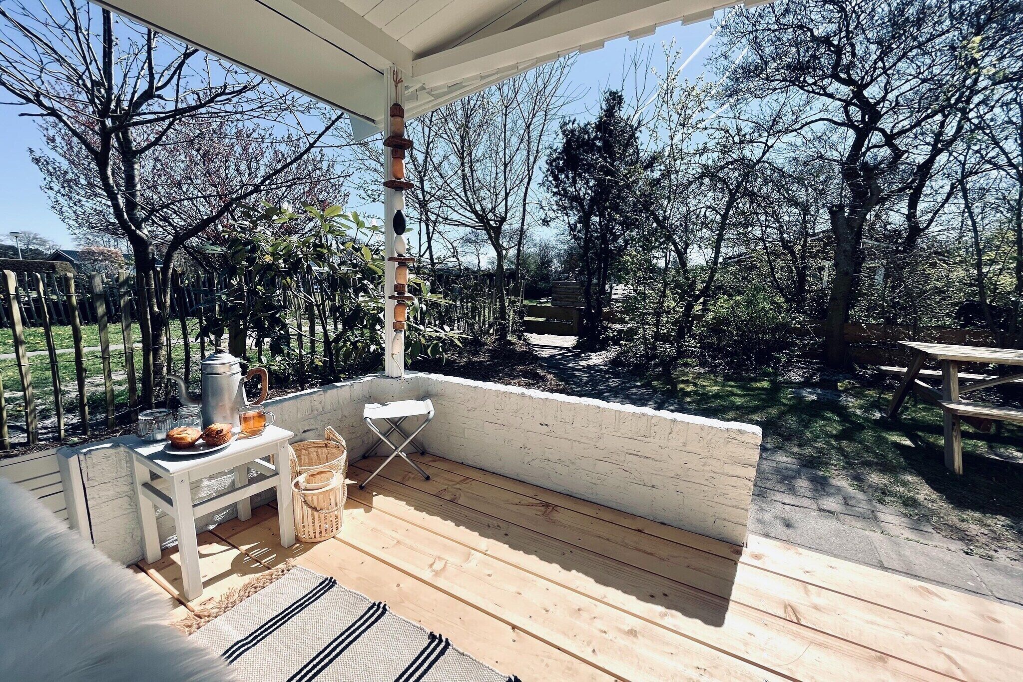 Een houten terras met een witte tafel en stoelen, een hangplant en een picknicktafel op de achtergrond.