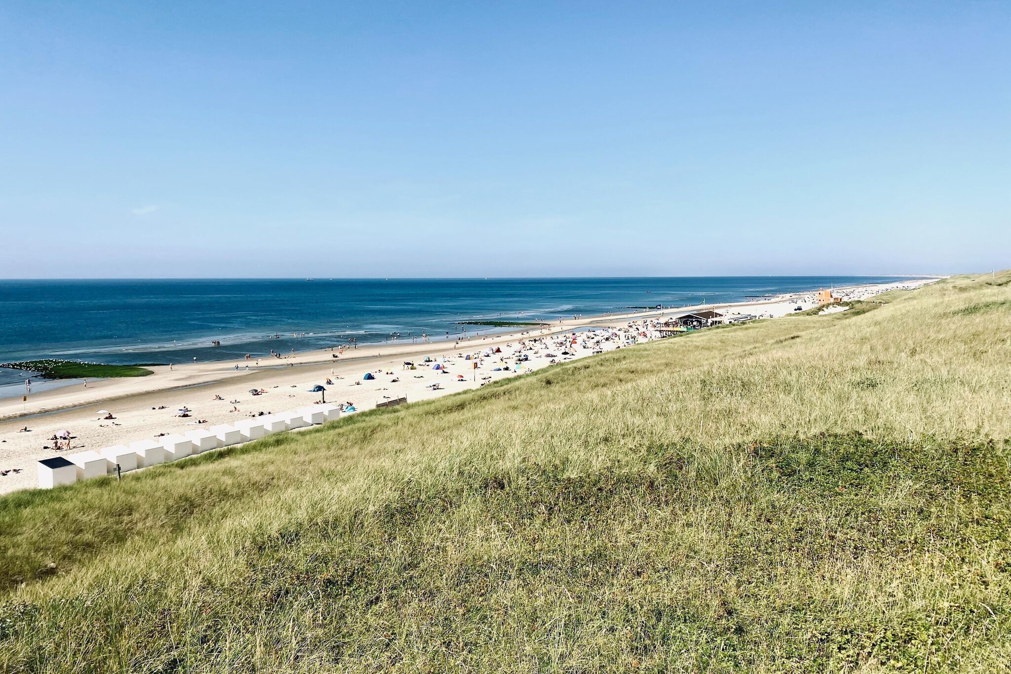 Ein Strand mit Zelten und Menschen, ein klarer Himmel und ein grasbewachsener Hügel.