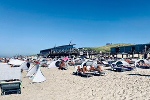 A beach with numerous tents and beach chairs, a large building with a stage, and a clear blue sky.