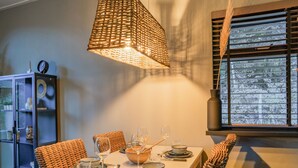 Un comedor con una mesa dispuesta para cuatro, una silla de mimbre, un otoman de mimbre, una cesta de mimbre y una lámpara colgante.