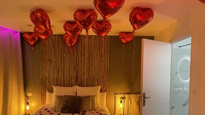A hotel room with a bed, rose petals, and heart-shaped balloons.