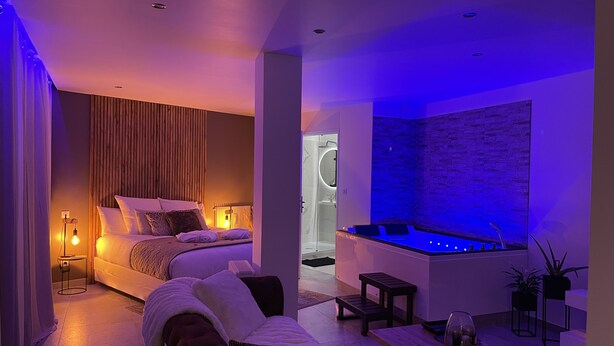 A modern bedroom with a bed, bedside table, and a small seating area. There is a bathroom with a bathtub and a shower. The room has a contemporary design with a neutral color palette.