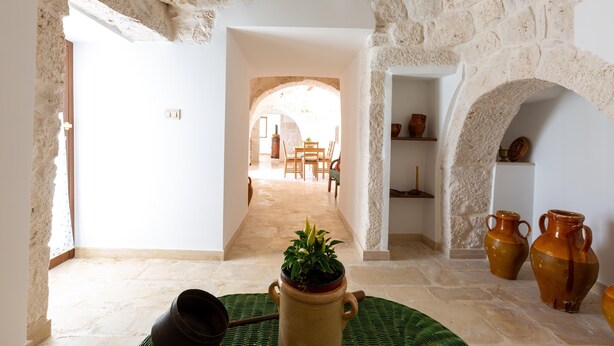 Una habitación con paredes de piedra, una planta en maceta, dos jarrones grandes y un tapete tejido en el suelo.