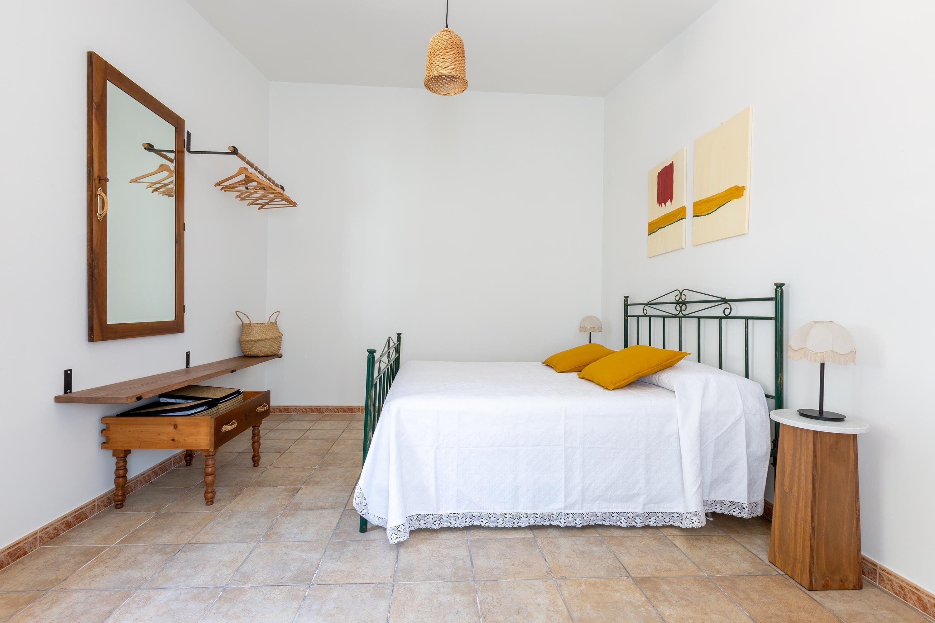 A bedroom with a bed, a wooden nightstand, a mirror, a hanging light fixture, and a wall-mounted shelf.