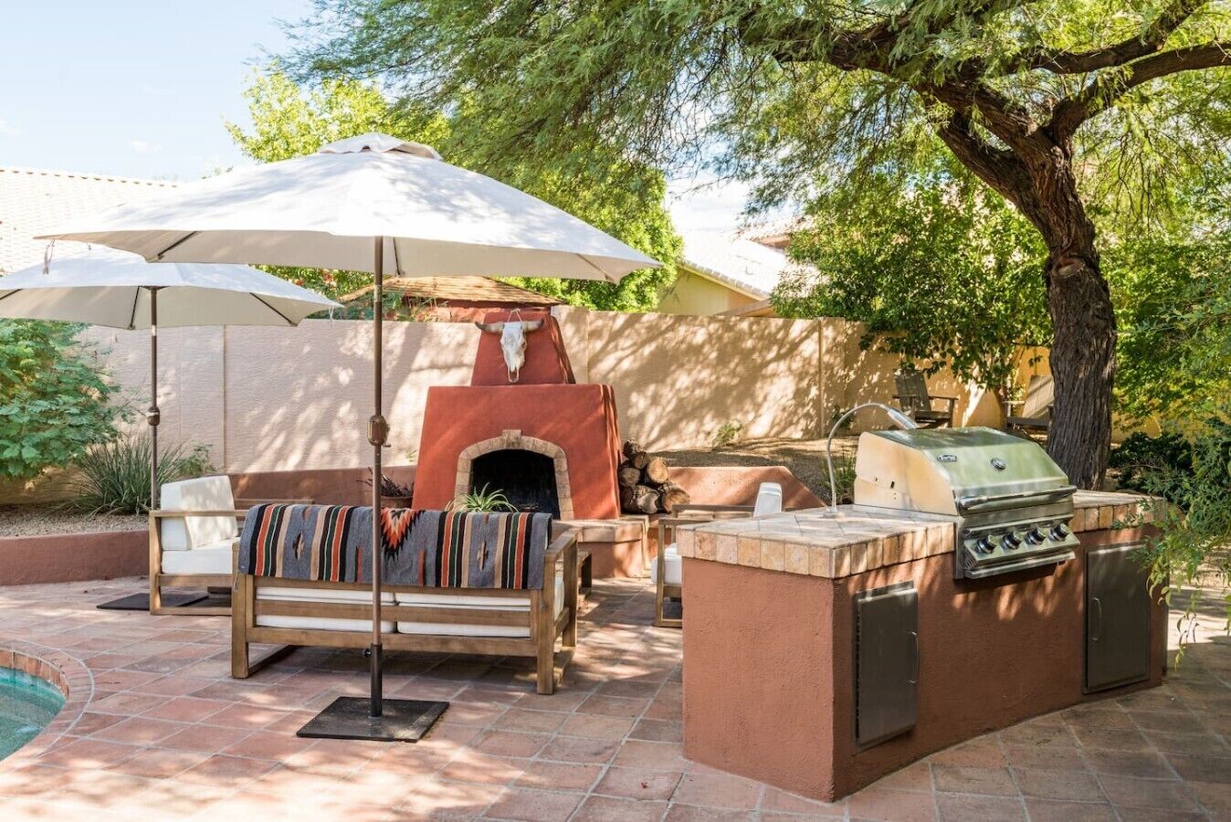 An outdoor patio with a stone barbecue, a wooden bench, and a large umbrella providing shade.