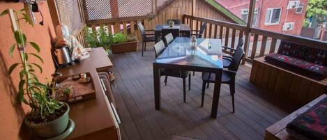 A wooden deck with a glass table and chairs, surrounded by potted plants and a wooden bench.