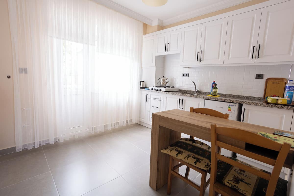 Una cocina moderna con armarios blancos, una mesa de comedor de madera y un ventanal con cortinas traslúcidas.