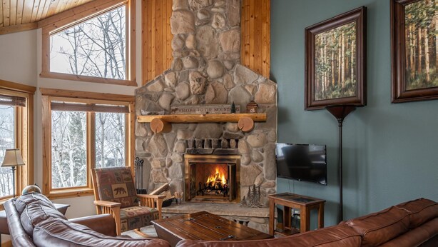 A cozy living room with a stone fireplace, leather seating, and large windows offering a view of trees.