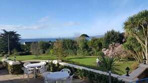 A patio with white chairs and tables, overlooking a well-maintained garden and a view of the sea.