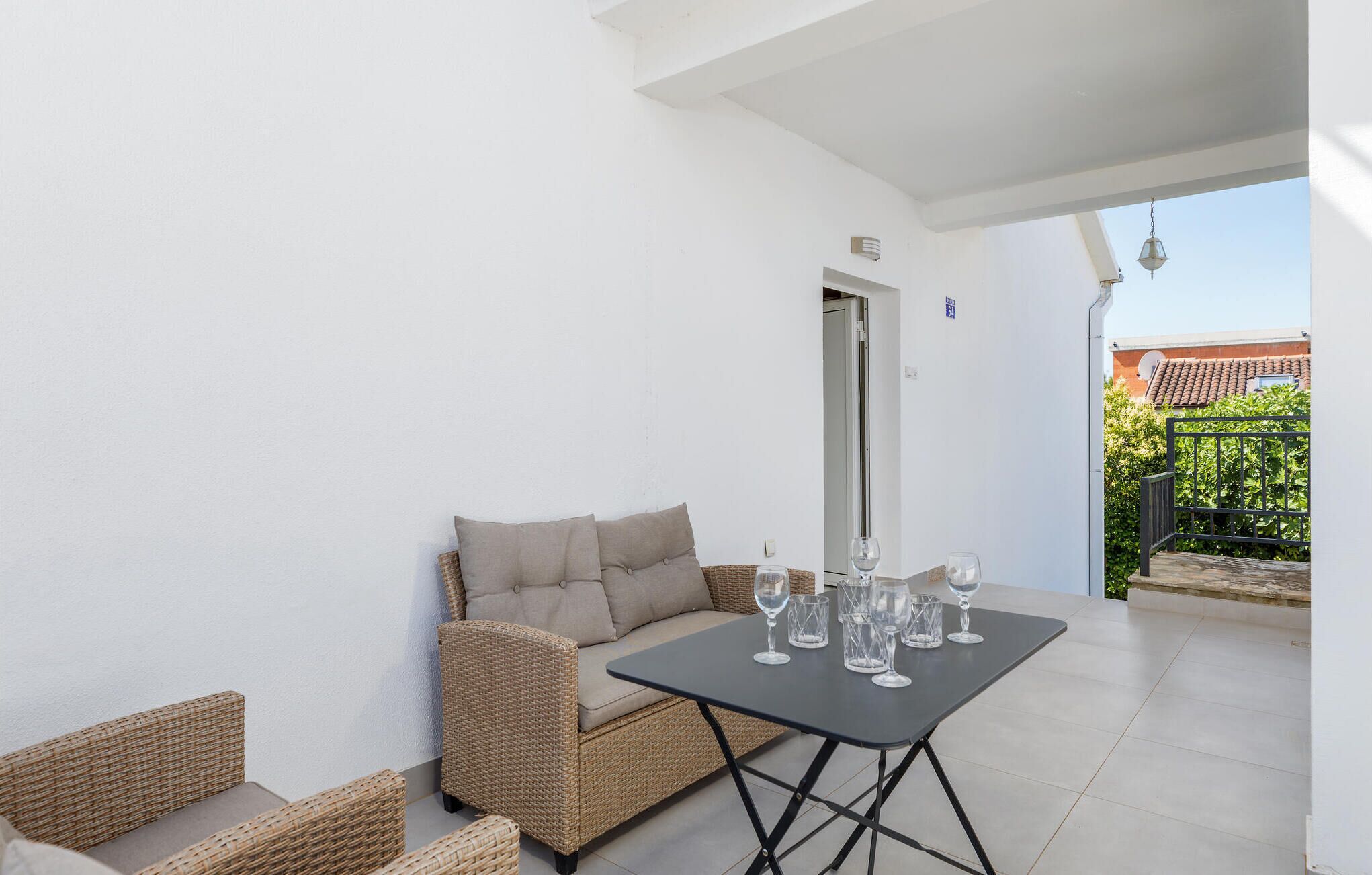 Une terrasse avec un canapé en osier, une petite table avec des verres à vin et une porte donnant sur un espace abrité.
