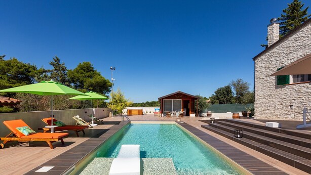 Una piscina con sillas de descanso y sombrillas, un edificio de piedra y una terraza de madera.