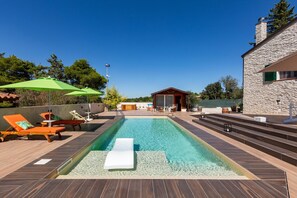 Una piscina con sillas de descanso y sombrillas, un edificio de piedra y una terraza de madera.