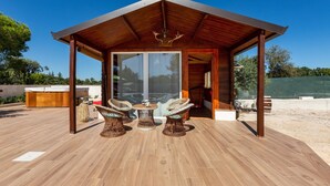 Un espacio de estar al aire libre cubierto, con piso de madera, sillas de mimbre y un vidrio que da al interior.