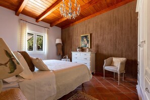 Un dormitorio con techo de madera, un candelabro, una cama con ropa de cama blanca, un aparador blanco, una silla y una ventana con cortinas.