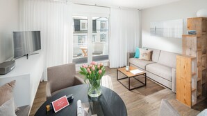 Una sala de estar moderna con un sofá, una mesa de centro y sillas. Un televisor está montado en la pared, y hay una ventana con cortinas.