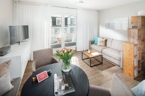 A modern living room with a sofa, coffee table, and chairs. A TV is mounted on the wall, and there is a window with curtains.