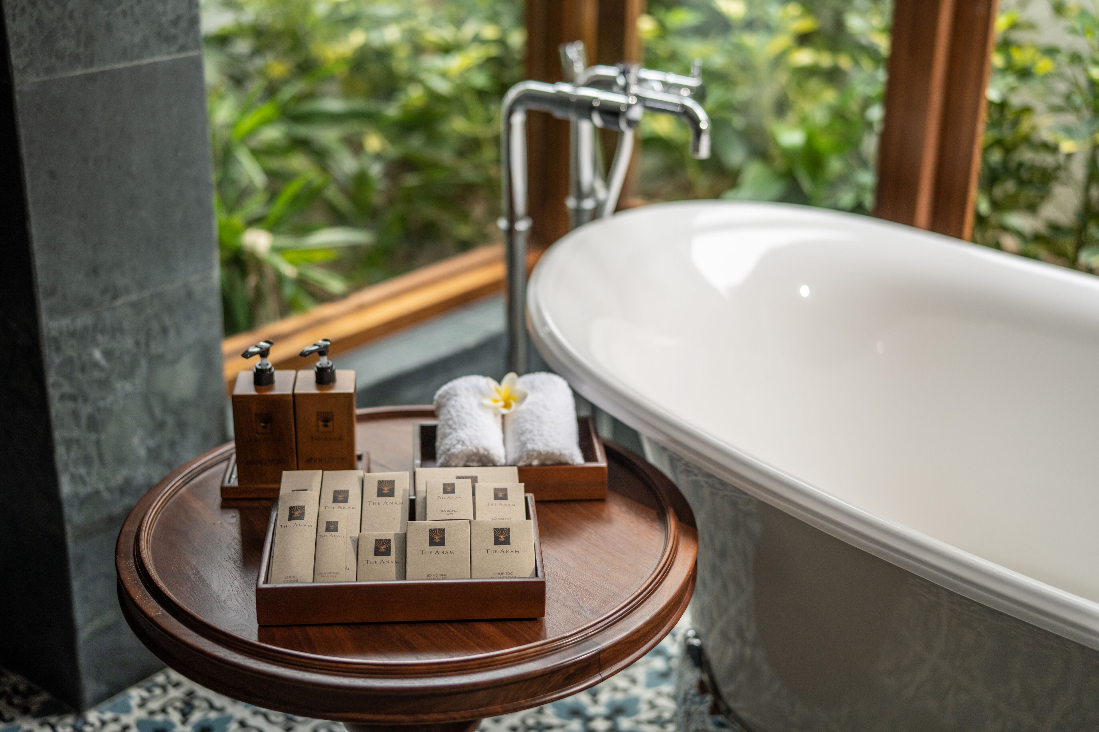 A bathroom with a bathtub, a wooden vanity with toiletries, and a view of greenery outside.