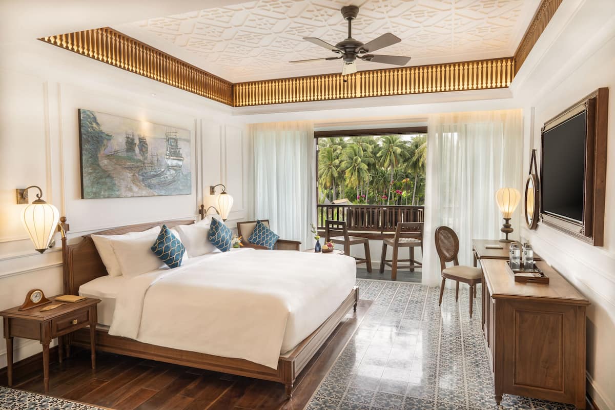 A spacious bedroom with a large bed, a ceiling fan, a painting, and a view of palm trees through the window.