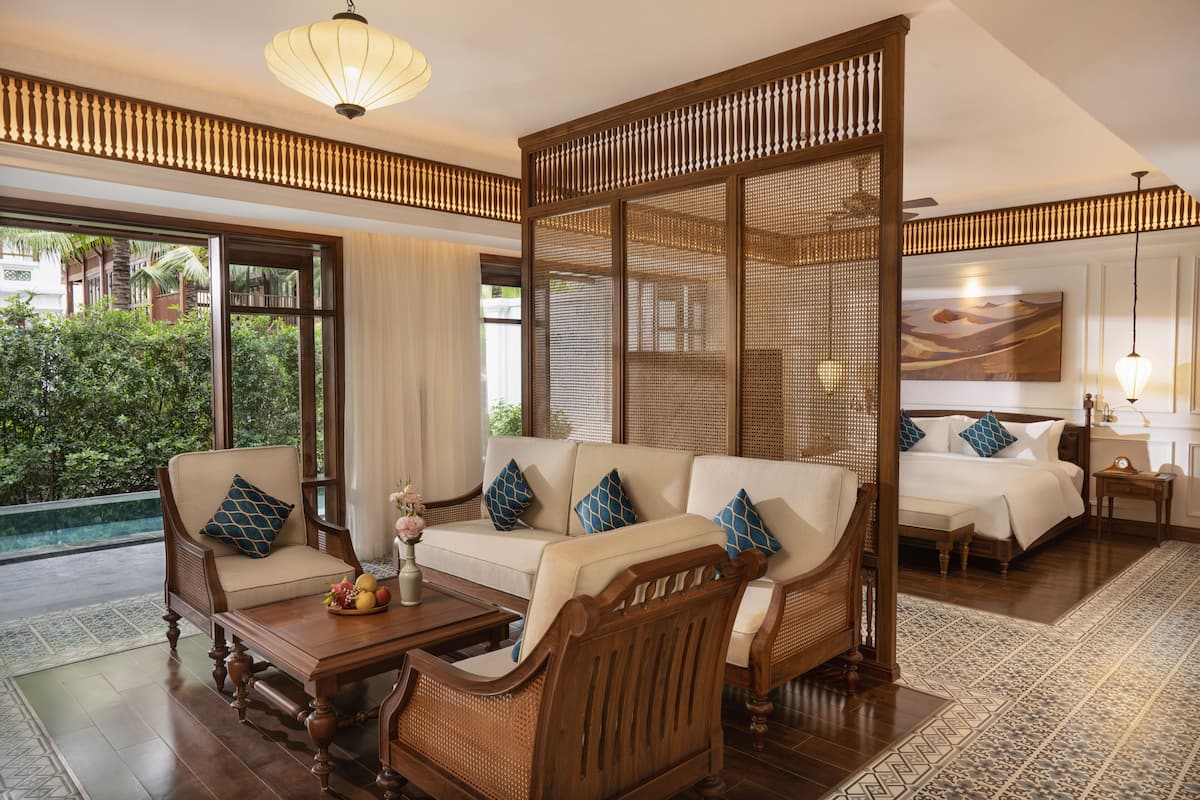 A room with a sofa, a wooden chair, a small table, and a bed. There is a window with a view of greenery outside.