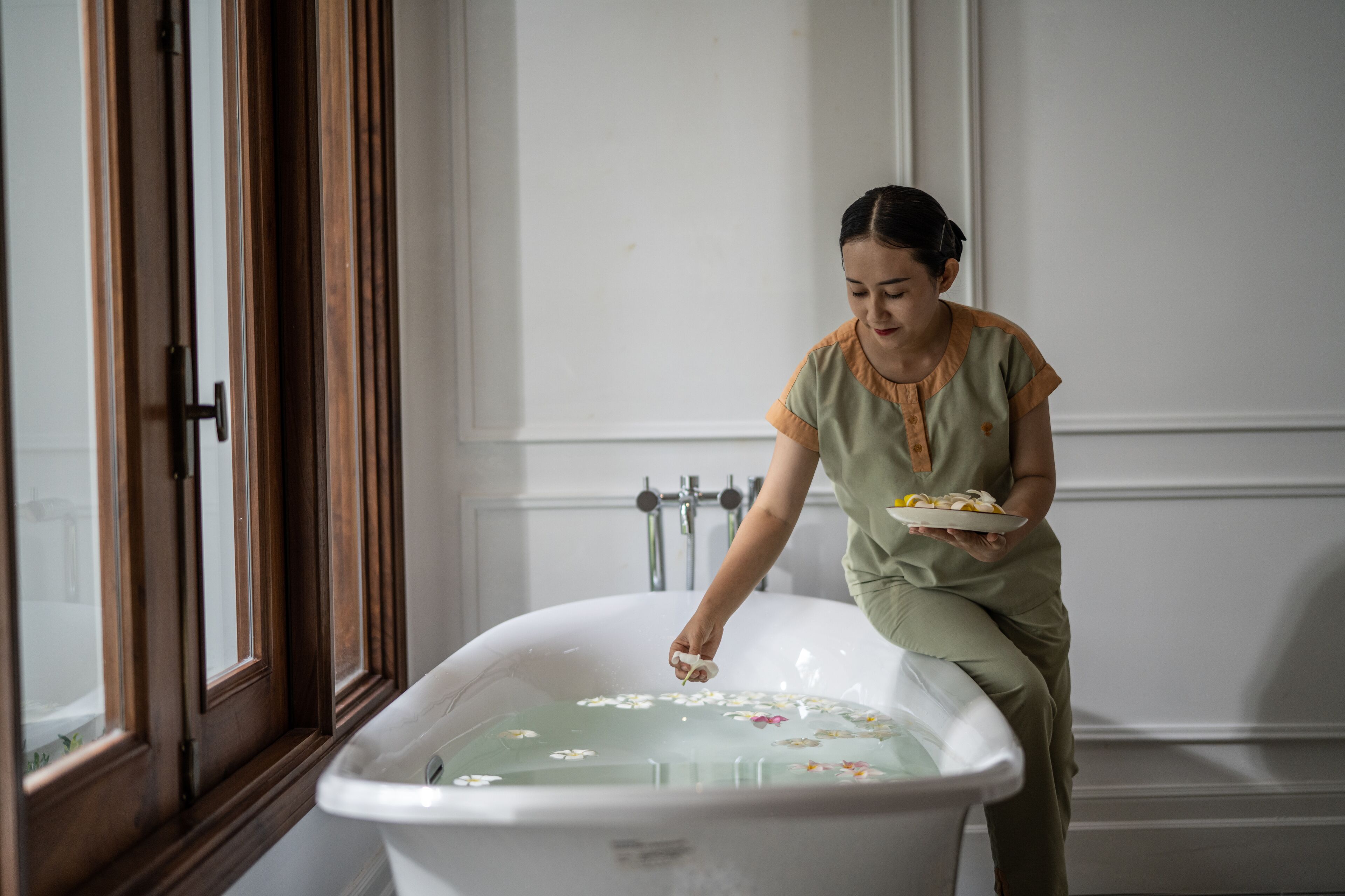 Eine Person in einem Badezimmer mit einer mit Wasser und Blumen gefüllten Badewanne, einem Fenster und einer Tür.