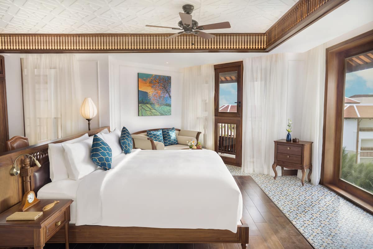 A spacious bedroom with a large bed, wooden headboard, a ceiling fan, a nightstand with a lamp, a small table with a vase, and a large window with a view.