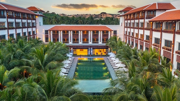 A resort with a central pool surrounded by palm trees, multiple buildings with balconies, and a mountainous backdrop. - The Anam Mui Ne (Mui Ne)