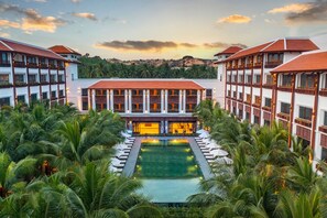 Un resort con una piscina central rodeada de palmeras, varios edificios con balcones y un paisaje montañoso al fondo.