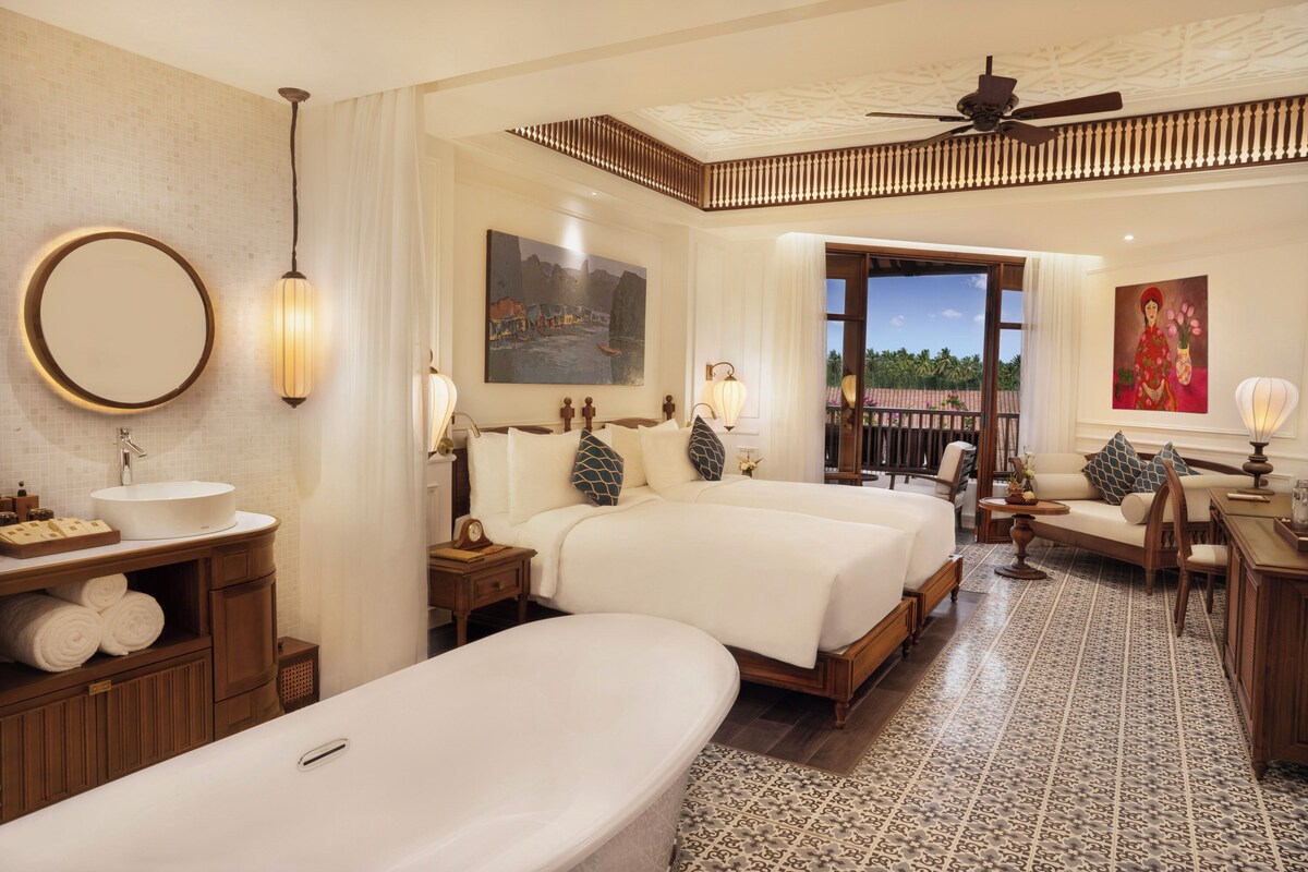 Habitación de hotel con una cama grande, un sofá independiente, una mesita, un escritorio, un espejo redondo, un ventilador de techo y vistas al exterior.