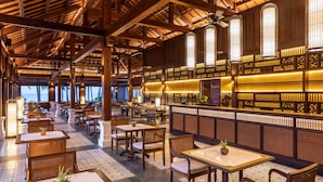 A spacious indoor dining area with wooden tables and chairs, a bar with decorative lighting, and a view of palm trees outside. - The Anam Mui Ne (Mui Ne)