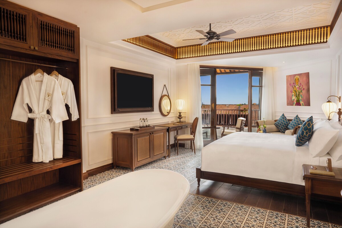 Habitación de hotel con una cama grande, una zona de estar con escritorio y silla, armario, televisión y balcón con vistas.