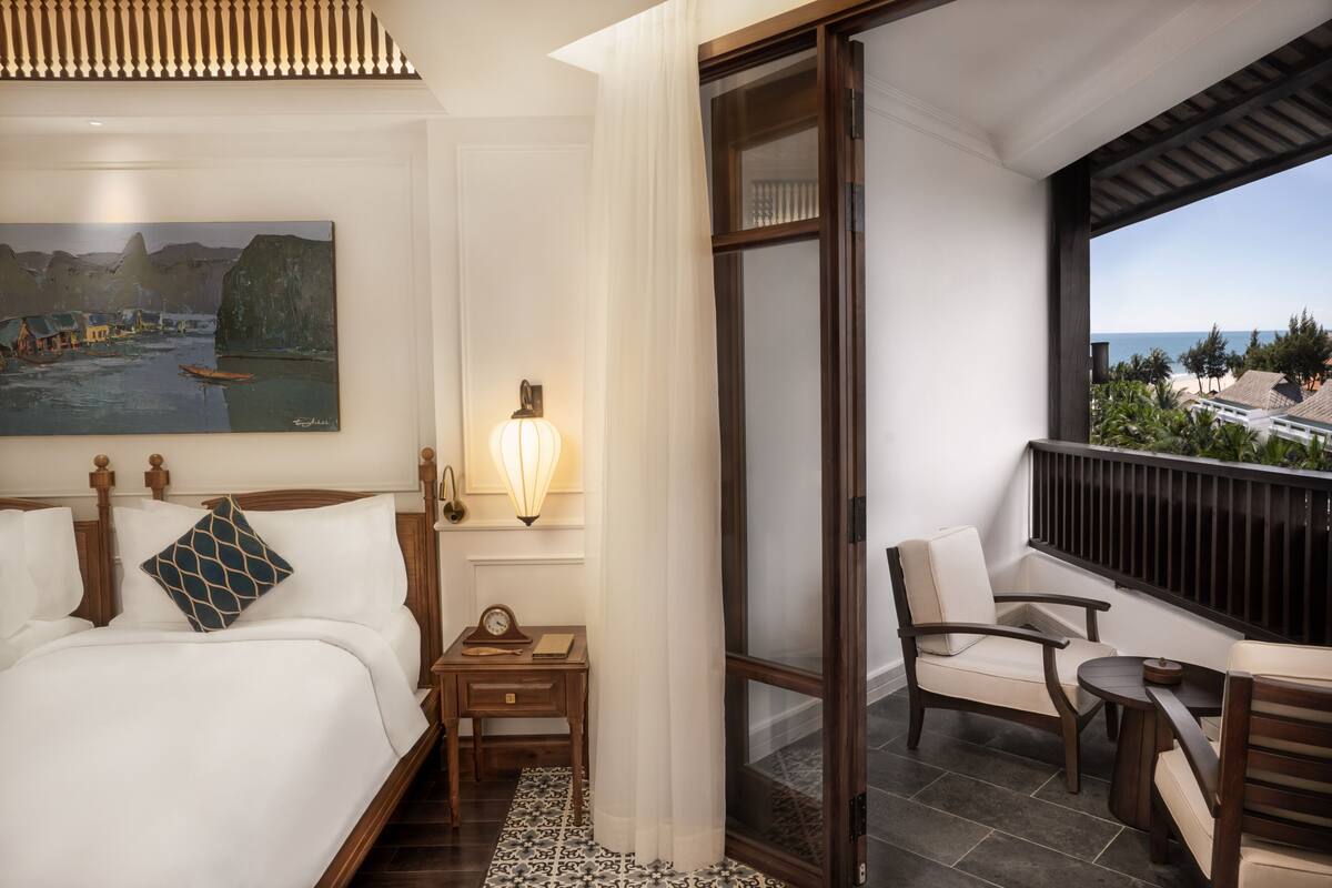 A hotel room with a bed, a nightstand, a lamp, a chair, and a balcony with a view of the beach and ocean.