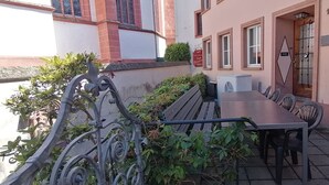 An outdoor dining area with a long table and chairs, surrounded by a wrought iron fence and greenery.