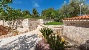 A stone pathway leading to a stone wall with a tiled roof, surrounded by greenery and trees. - 2061 Villa Trulli Natura by Perle di Puglia (Noci)
